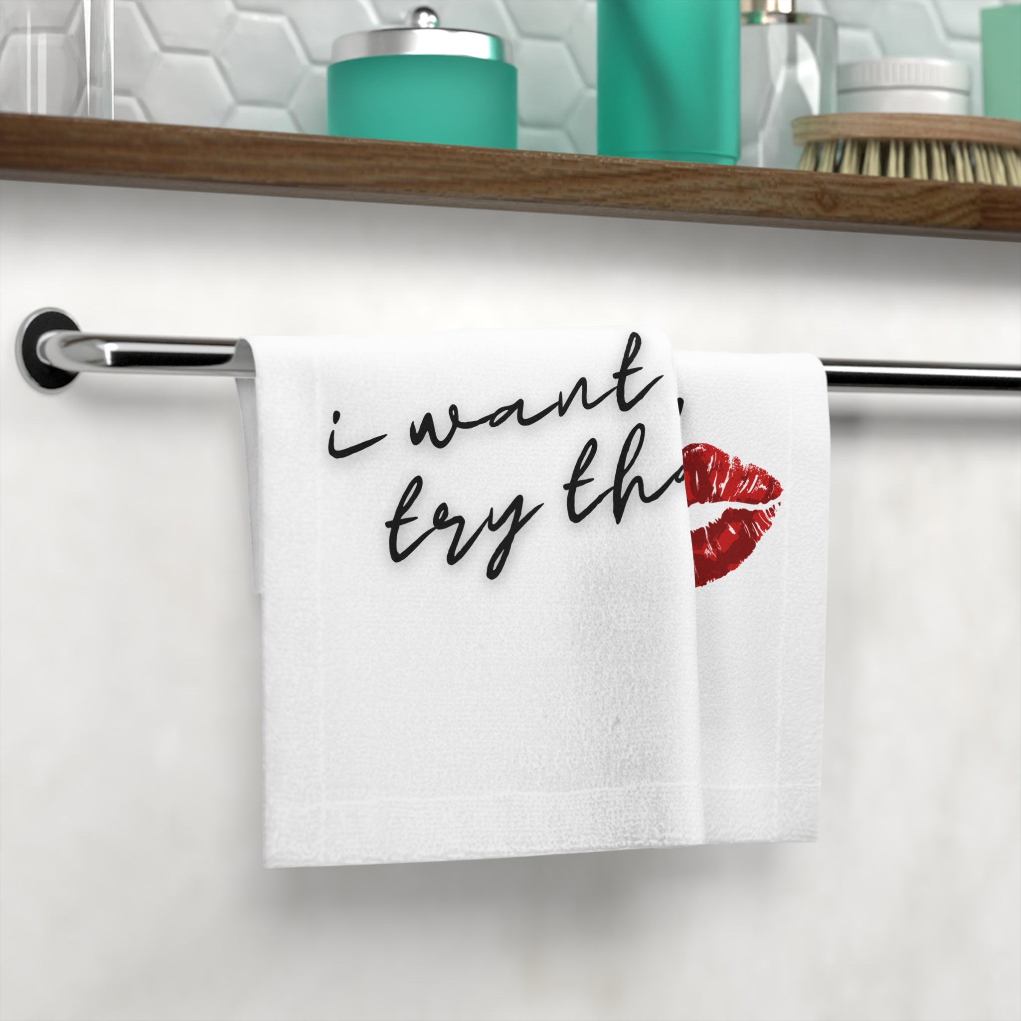 Decorative white hand towel with 'I want to try that!' text and red lipstick kiss, displayed on a bathroom towel rack.